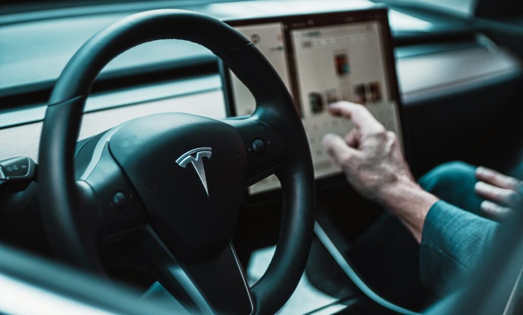 Czarne chmury nad Teslą Wnętrze samochodu Tesla. Kierownica z logo firmy i dłoń mężczyzny skierowana na komputer pokładowy.