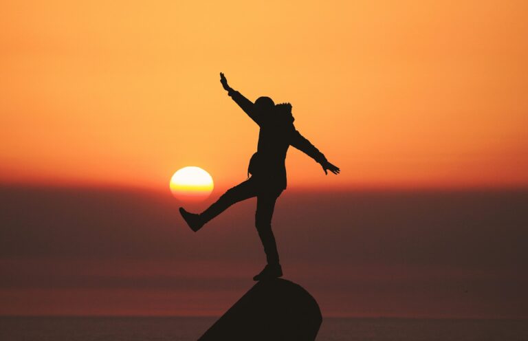 Mężczyzna stojący na szczycie o zachodzie słońca. Jedną nogę unosi w powietrzu, sprawając wrażenie jakby podtrzymywał nią kulę słońca.